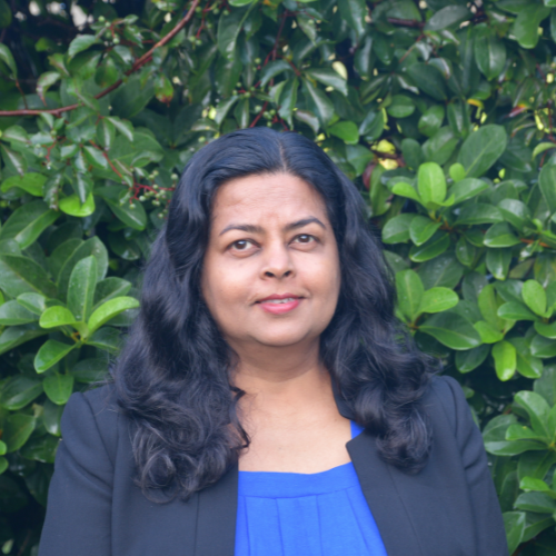 Dr. Uttara Samarakoon in a dark blazer and blue top in front of green foliage.