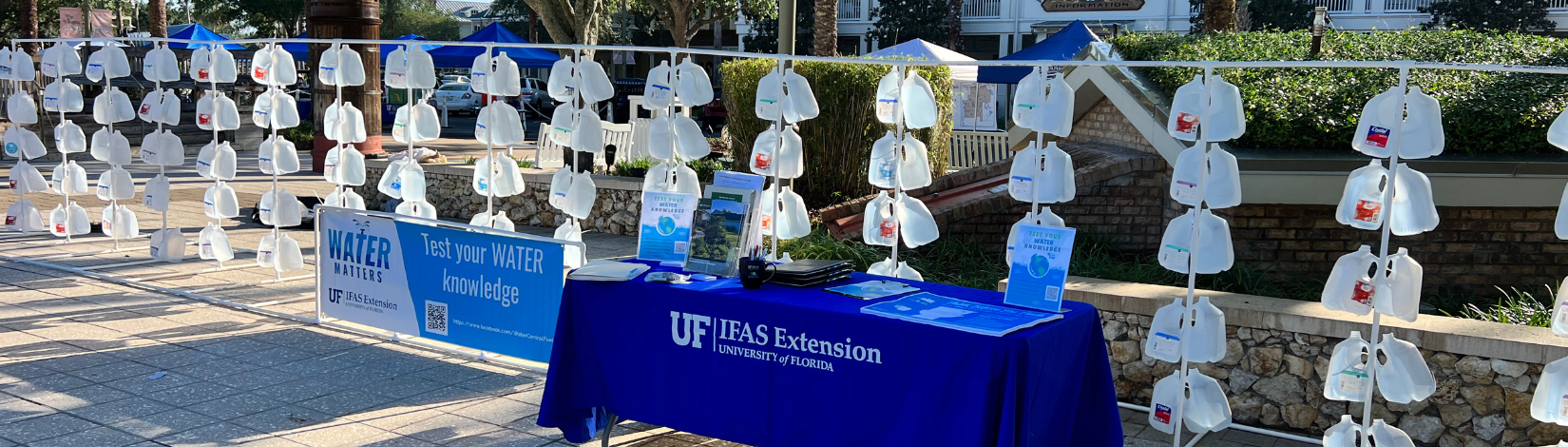 Outdoor educational display with hanging gallon jugs and a UF IFAS Extension table.
